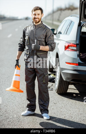 Portrait Of Happy Pannenhilfe Arbeiter in Uniform stehend mit Kegel in der Nähe des Auto auf den Straßenrand Stockfoto