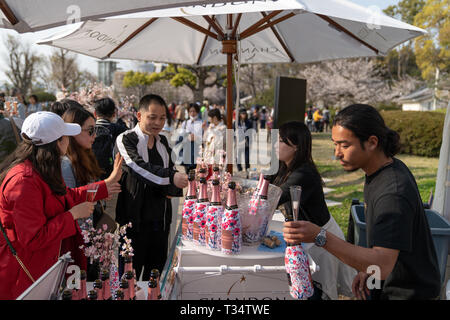 Cherry Blossom Festival in der Burg von Osaka. Champagner mit weißen Sonnenschirm und Thekenbereich mit mehreren Flasche Champagner stall in Papier mit Kirschblüten auf gewickelt. Menschen kaufen Gläser Champagner von zwei Menschen behidn der Stall. Stockfoto