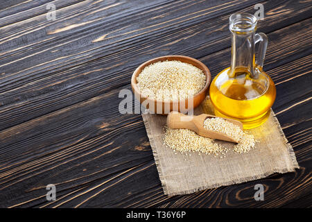 Sesamöl in Glasflasche und Samen in der Schüssel mit Schaufel auf dunklen Holztisch Stockfoto