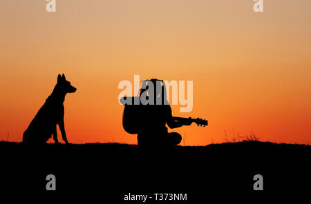 Silhouettegirls Gitarrist auf Sonnenuntergang Hintergrund, Silhouette einer Hunderasse Belgischer Schäferhund Malinois, glücklich, Freunde, outdoor Stockfoto