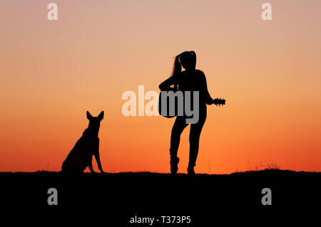 Silhouettegirls Gitarrist auf Sonnenuntergang Hintergrund, Silhouette einer Hunderasse Belgischer Schäferhund Malinois, glücklich, Freunde, outdoor Stockfoto