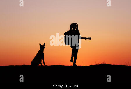 Silhouettegirls Gitarrist auf Sonnenuntergang Hintergrund, Silhouette einer Hunderasse Belgischer Schäferhund Malinois, glücklich, Freunde, outdoor Stockfoto