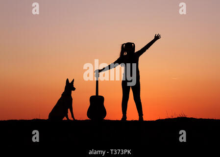 Silhouettegirls Gitarrist auf Sonnenuntergang Hintergrund, Silhouette einer Hunderasse Belgischer Schäferhund Malinois, glücklich, Freunde, outdoor Stockfoto