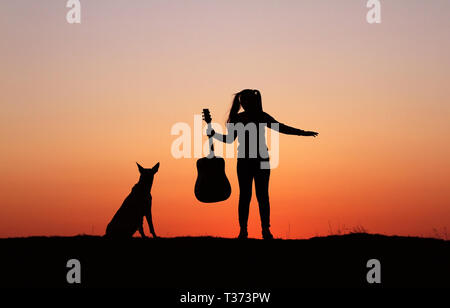 Silhouettegirls Gitarrist auf Sonnenuntergang Hintergrund, Silhouette einer Hunderasse Belgischer Schäferhund Malinois, glücklich, Freunde, outdoor Stockfoto