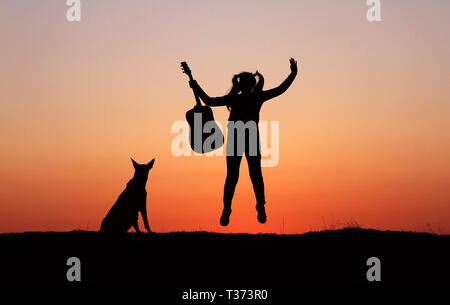 Silhouettegirls Gitarrist auf Sonnenuntergang Hintergrund, Silhouette einer Hunderasse Belgischer Schäferhund Malinois, glücklich, Freunde, outdoor Stockfoto