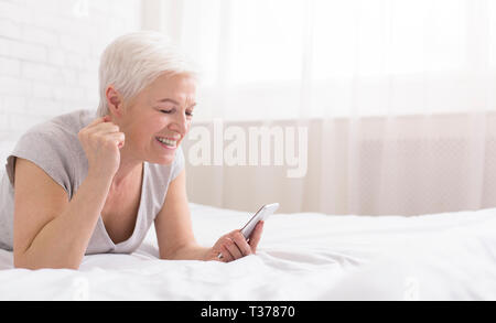 Gute Nachrichten. Gerne ältere Frau auf dem Bett liegend, lächelnd und Lesen von SMS auf Handy, genießen gewinnen, Panorama mit leeren Raum Stockfoto