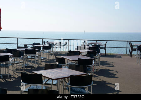 Leere Cafe im frühen Frühling am Bodensee in Deutschland, Tischen und Stühlen ohne Personen an der Seepromenade gegen Bodensee und Rauszuschwimmen Stockfoto