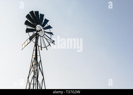 Low Angle View eines altmodischen, multi-Klinge, Metall wind Pumpe auf einem gittermast in der Hintergrundbeleuchtung gegen einen hellblauen Himmel. Stockfoto