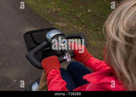 Ein über die Schulter geschossen von einer älteren Dame in einem roten Mantel, in der Freiheit eines elektrischen Mobilität scooter, ein Drittel der Kopf, Stockfoto