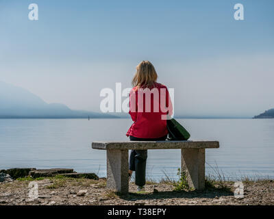 Bild der Frau mit roter Jacke sitzt auf einer Steinbank am Lago Maggiore mit Berge und Wolken suchen Stockfoto