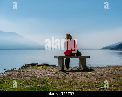 Bild der Frau mit roter Jacke sitzt auf einer Steinbank am Lago Maggiore mit Berge und Wolken suchen Stockfoto