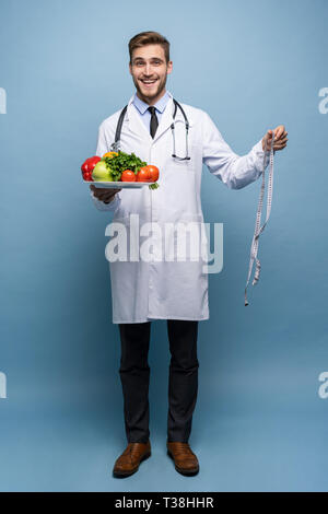 Arzt mit einem Maßband und einem Teller mit Gemüse und Obst isoliert auf hellblauem Hintergrund. Stockfoto