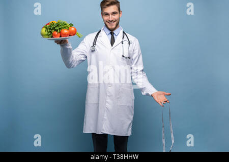 Arzt mit einem Maßband und einem Teller mit Gemüse und Obst isoliert auf hellblauem Hintergrund. Stockfoto