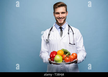 Arzt suchen an Kamera und halten Teller mit frischem Gemüse auf Hellblau, gesunde Ernährung Konzept isoliert. Stockfoto