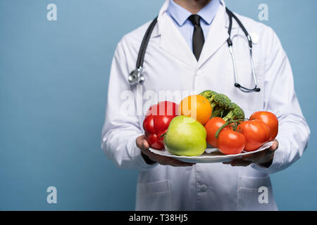 Arzt hält Teller mit frischem Gemüse auf Hellblau, gesunde Ernährung Konzept isoliert. Stockfoto