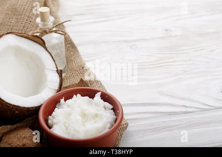 Kokosöl in braun Ton Schale, Glas, Flasche, Kokosnussschalen mit Fleisch auf Hanf Sack auf weißen Küche aus Holz Tisch Stockfoto
