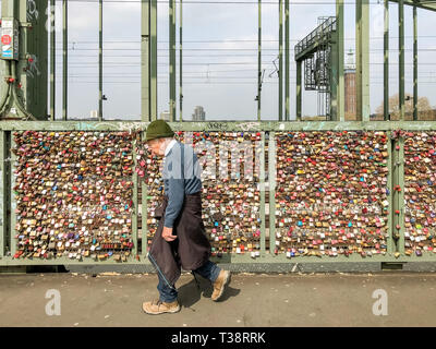 Köln, Deutschland, 6. April 2019. Alter Mann vorbei vor der unzähligen bunten Vorhängeschlösser von Liebhabern auf einem Zaun auf der Hohenzollern Brücke links Stockfoto
