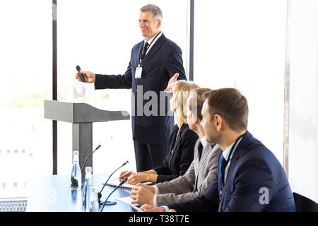Öffentliche Lautsprecher mit Mikrofon auf der Pressekonferenz sprechen hinter der Tribüne, seine Kollegen am Tisch Stockfoto