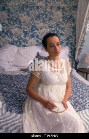 Attraktive junge Frau in authentischen Regency Kleid sitzt auf einem antiken Bett Stockfoto