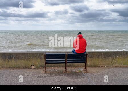 Männliche Figur im roten Mantel sitzt auf der Rückseite der Rücksitzbank aus Blick auf das Meer an der Küste von Hove, East Sussex England Großbritannien Stockfoto