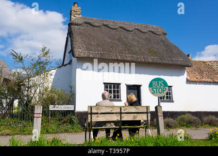 Thriplow, Cambridge, England, UK - März 2019: Älteres Ehepaar sitzt auf einer Holzbank an einem Dorf Bushaltestelle mit traditionellen britischen Cottage Stockfoto