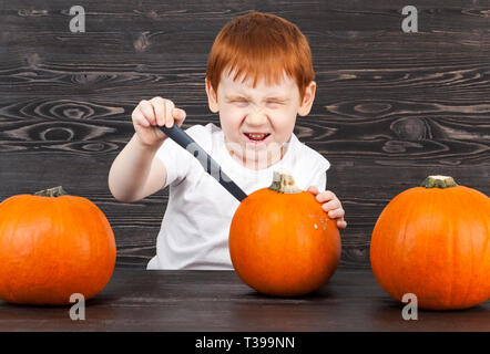 Rothaarige Junge mit roten Kürbisse in Vorbereitung für Halloween, Schnitte Kürbis mit Aufwand und Emotionen Stockfoto