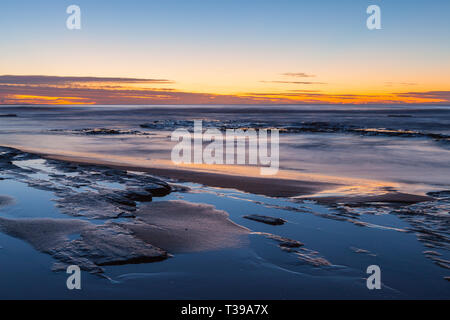 Küsten lange Belichtung um Sonnenaufgang mit Strand, Felsen und Wasser im Vordergrund und Horizont mit Wolken im Himmel im Hintergrund Stockfoto