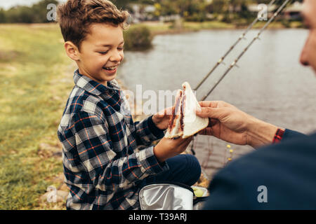 Kind eine Pause vom Angeln Sandwiches zu essen. Mann, Sandwich zu seinem Kind, während neben einem See zum Angeln zu sitzen. Stockfoto