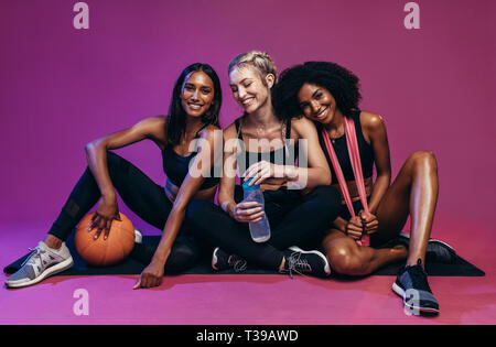 Gruppe von drei fitness Frauen Ausruhen nach Training. Multi-ethnischen Gruppe von Frauen im Sport Kleidung sitzen auf farbigen Hintergrund mit Basketball, wat Stockfoto
