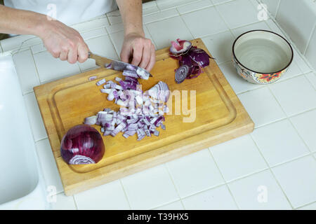 Ansicht von oben der Arbeitsplatte in der Küche, ein Schneidbrett, Schüssel mit Wasser, Ecke von Spülbecken, mit weiblichen Händen würfeln rote Zwiebel. Stockfoto