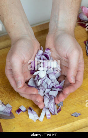 Nahaufnahme, Ansicht von oben von zwei weiblichen Händen schröpfen gewürfelte rote Zwiebel auf einem Schneidebrett statt. Stockfoto