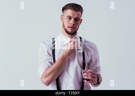 Eleganten und stilvollen Mann in der klassischen Verschleiß holding Glas mit wiskey in der einen Hand und Stellring mit einem anderen beim Stehen gegen Grau backgrou Stockfoto