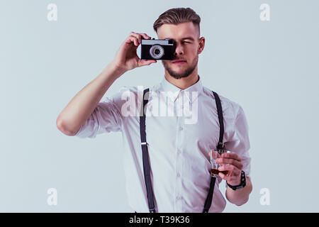 Schöne und stilvolle Mann im weißen Hemd und Hosenträgern, Foto auf alten Vintage Kamera und halten Glas Whisky in der Hand, während gegen Ständigen Stockfoto