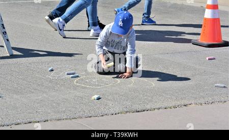 Junger ethnischer Junge, der Kreidezeichnung auf der Straße macht Ein Parkplatz, wenn die Leute mit Spaß laufen Eine Mütze Stockfoto