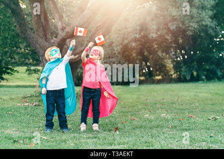 Kaukasische Kinder in Superhelden Kostüme und Masken Holding winken kanadischen Flagge. Jungen, Mädchen feiert Nationalfeiertag Kanada Tag im Sommer, Park Stockfoto