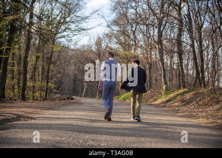 Rückansicht, zwei homosexuelle Menschen auf dem Weg in den Wald, auf Asphalt. Zusammen halten sich an den Händen. Stockfoto