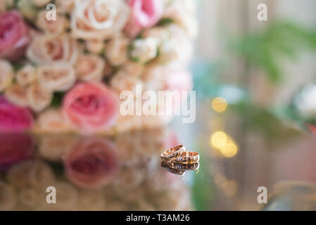 Schöne goldene Hochzeit Ringe auf der Glasoberfläche mit Reflexion auf dem Hintergrund der Blumenstrauß für die Braut von Rosen Stockfoto