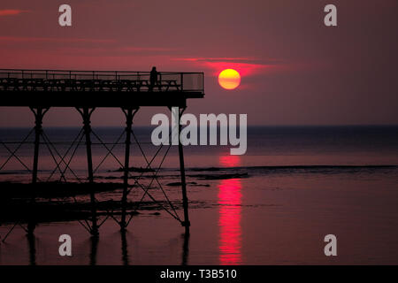 Aberystwyth, Wales, UK. 8. Apr 2019. Nach einem kühlen, grau, bewölkten Tag, die Sonne macht eine kurze, aber spektakulären Auftritt, Sie hinter der markanten Silhouette der Küste pier, kurz vor Sonnenuntergang, in Aberystwyth, auf der Cardigan Bay Küste von West Wales. Credit: Keith Morris/Alamy leben Nachrichten Stockfoto