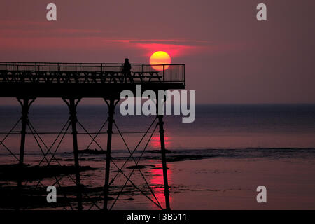 Aberystwyth, Wales, UK. 8. Apr 2019. Nach einem kühlen, grau, bewölkten Tag, die Sonne macht eine kurze, aber spektakulären Auftritt, Sie hinter der markanten Silhouette der Küste pier, kurz vor Sonnenuntergang, in Aberystwyth, auf der Cardigan Bay Küste von West Wales. Credit: Keith Morris/Alamy leben Nachrichten Stockfoto