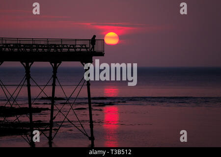 Aberystwyth, Wales, UK. 8. Apr 2019. Nach einem kühlen, grau, bewölkten Tag, die Sonne macht eine kurze, aber spektakulären Auftritt, Sie hinter der markanten Silhouette der Küste pier, kurz vor Sonnenuntergang, in Aberystwyth, auf der Cardigan Bay Küste von West Wales. Credit: Keith Morris/Alamy leben Nachrichten Stockfoto