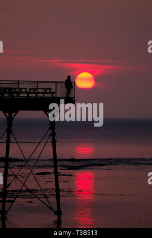 Aberystwyth, Wales, UK. 8. Apr 2019. Nach einem kühlen, grau, bewölkten Tag, die Sonne macht eine kurze, aber spektakulären Auftritt, Sie hinter der markanten Silhouette der Küste pier, kurz vor Sonnenuntergang, in Aberystwyth, auf der Cardigan Bay Küste von West Wales. Credit: Keith Morris/Alamy leben Nachrichten Stockfoto