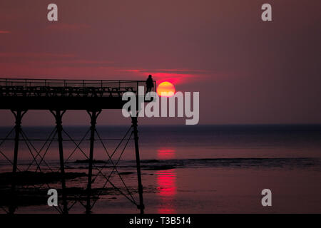 Aberystwyth, Wales, UK. 8. Apr 2019. Nach einem kühlen, grau, bewölkten Tag, die Sonne macht eine kurze, aber spektakulären Auftritt, Sie hinter der markanten Silhouette der Küste pier, kurz vor Sonnenuntergang, in Aberystwyth, auf der Cardigan Bay Küste von West Wales. Credit: Keith Morris/Alamy leben Nachrichten Stockfoto