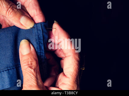 Nahaufnahme einer Hand der alten Frau mit Gewinde mit der Nadel in der Hose mit schwarzem Hintergrund Stockfoto