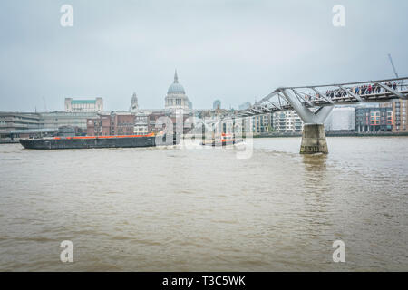 An einem nebligen Tag in London, Großbritannien, führt ein großer Lastkahn, der von einem Schlepper gezogen wird, unter der Millennium Bridge hindurch Stockfoto