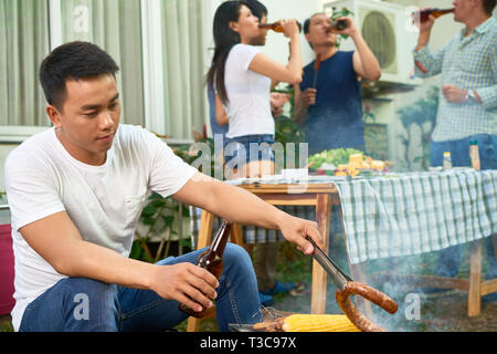 Vietnamesische jungen Mann kochen Wurst vom Grill, wenn seine Freunde sprechen im Hintergrund Stockfoto