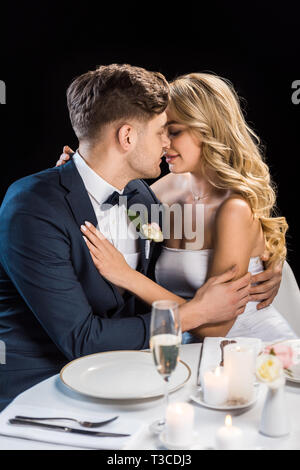 Brautpaar umarmen beim Sitzen am Tisch serviert auf Schwarz isoliert Stockfoto