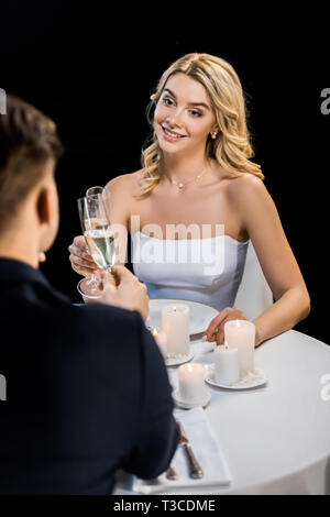 Selektiver Fokus der junge Paar klirren Gläser Champagner beim Sitzen am Tisch serviert auf Schwarz isoliert Stockfoto