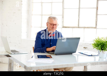 Portrait von älteren Geschäftsmann mit grauen Haaren sitzen im Büro und arbeitet an seinem Laptop. Ältere professionelle Mann mit Hemd und Fliege. Stockfoto