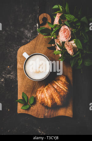 Kaffeepause. Flachbild-lay von Tasse heißen Cappuccino und Croissant und Bouquet von rosa Blüten über schwarzen schäbig Hintergrund, Ansicht von oben, vertikale compositio Stockfoto