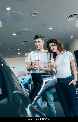 Fröhlicher Mann hält Papier Cup in der Nähe von Curly stehende Frau mit Hand in der Tasche, während im Automobil suchen Stockfoto